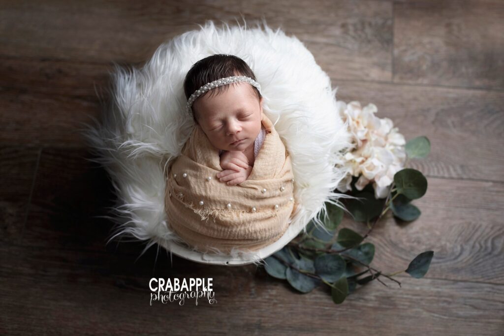 A stylish and neutral newborn photo of a baby girl wrapped in a light swaddle embellished with pearls, wearing a matching pearl headband. She is on a white fur, on top of a dark rustic wooden background. The portrait is accessorized with faux eucalyptus leaves.