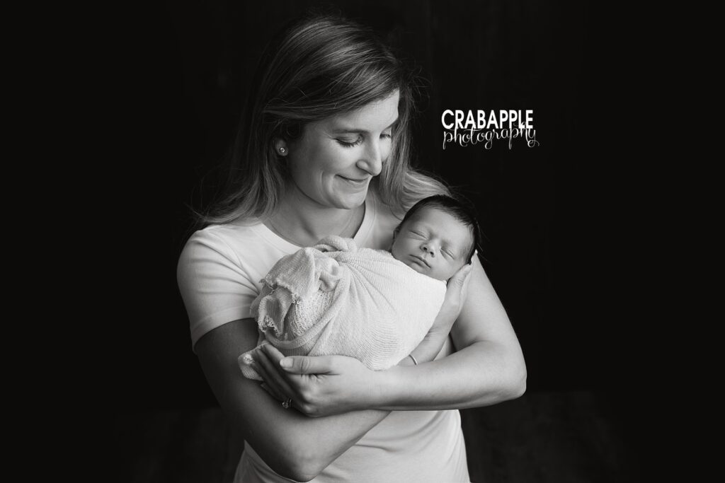 Black and white portrait of mother and newborn. Mom is in front of a black background and smiling down at her daughter in her arms.