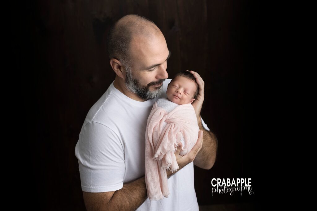 Portrait of father and newborn. He is in a white tee shirt in front of a black background and looking down at his daughter wrapped in pink.