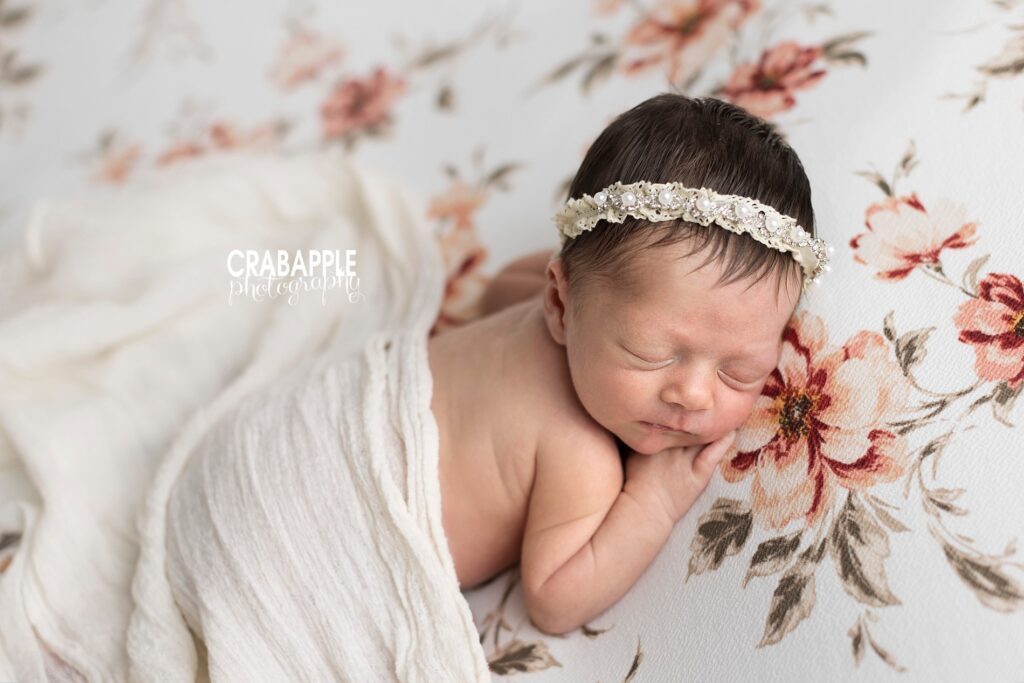 Newborn portrait of a sleeping baby girl draped with a white wrap on top of a pink floral blanket. She is wearing a dainty pearl and lace headband.