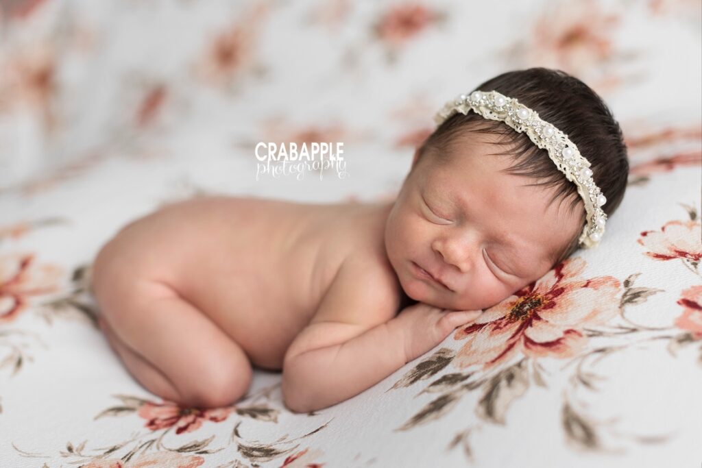 Sleeping newborn photographed on a white blanket with pink flowers. She is wearing a classic pearl and lace headband.