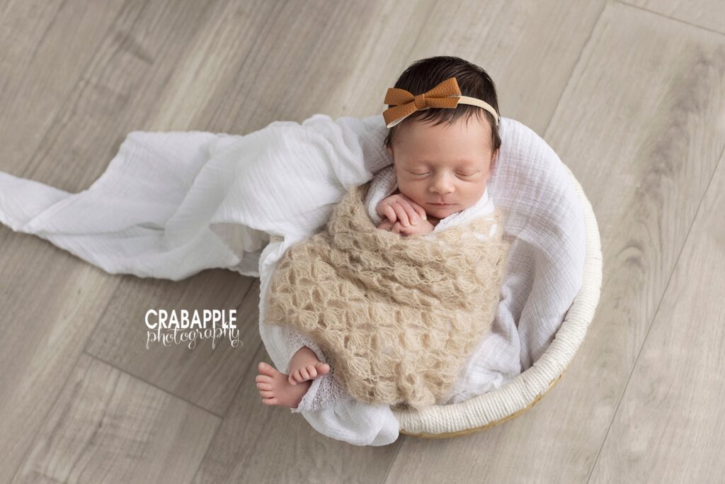 Neutral newborn photo of a sleeping baby girl wrapped in a knit beige blanket. She is laying on her back inside of a basket draped with white blanket. The backdrop is a rustic white washed wooden floor. She is wearing a brown leather bow headband.