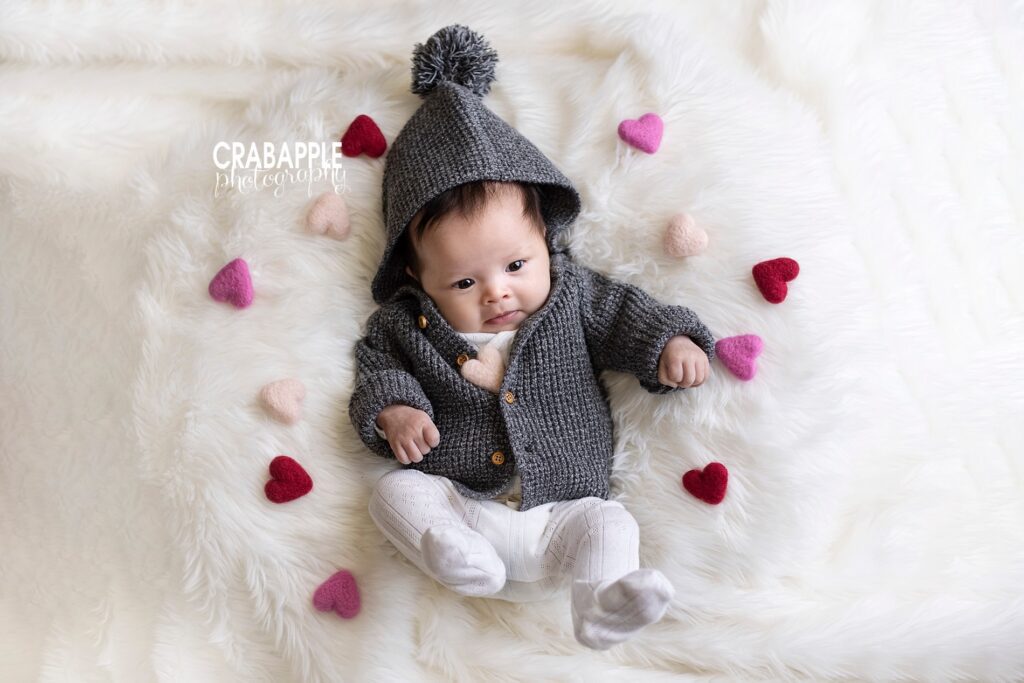 Photo of a 3 month old baby girl wearing a dark gray knit sweater with a pom-pom, on a white fur, surrounded by red and pink felt hearts.