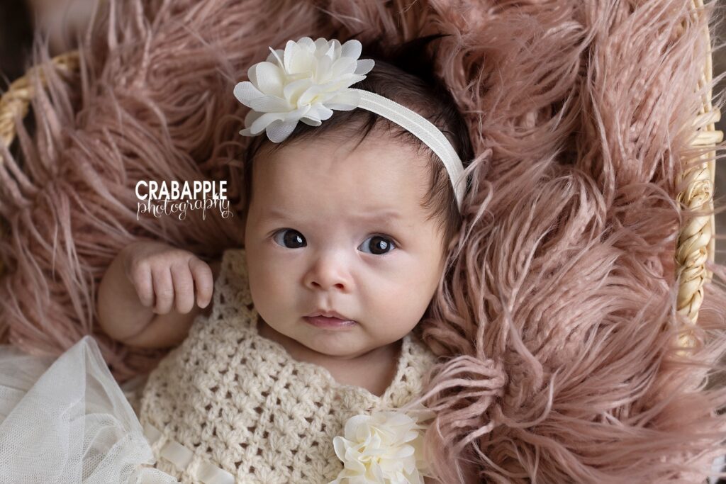 Close up portrait of a 3 month old baby girl with brown hair and eyes wearing a white flower headband and laying on a light pink fur.