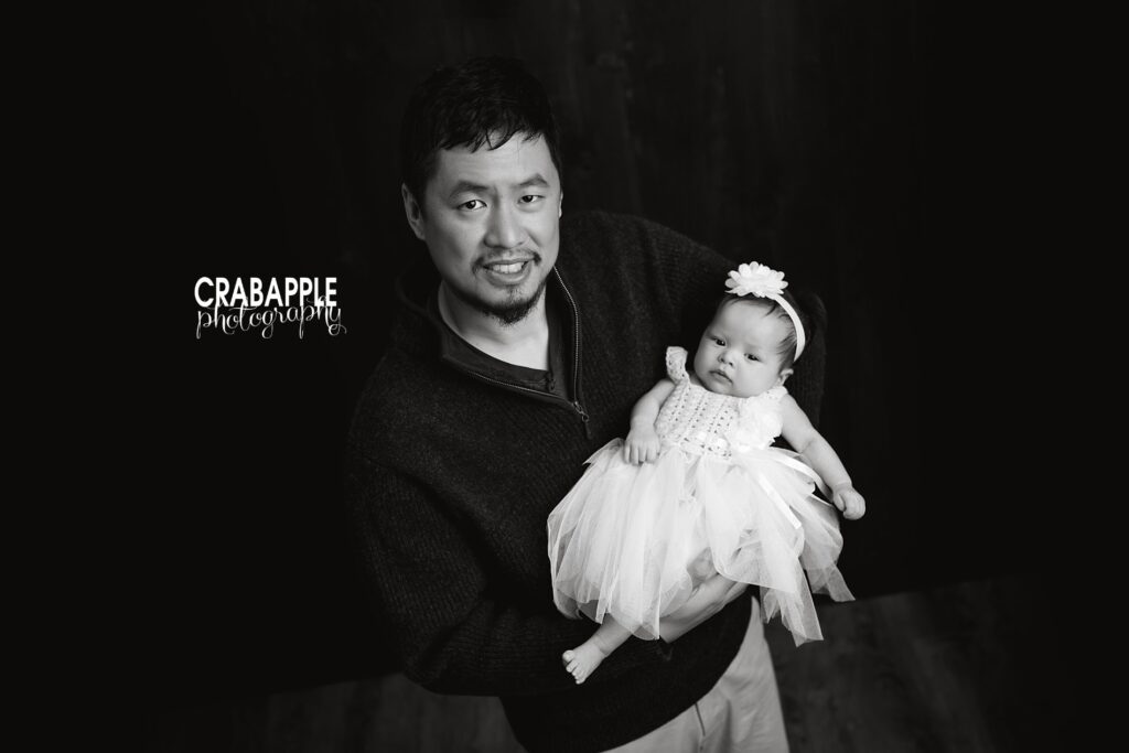 A black and white portrait of a father standing in front of a dark background holding his three month old daughter.