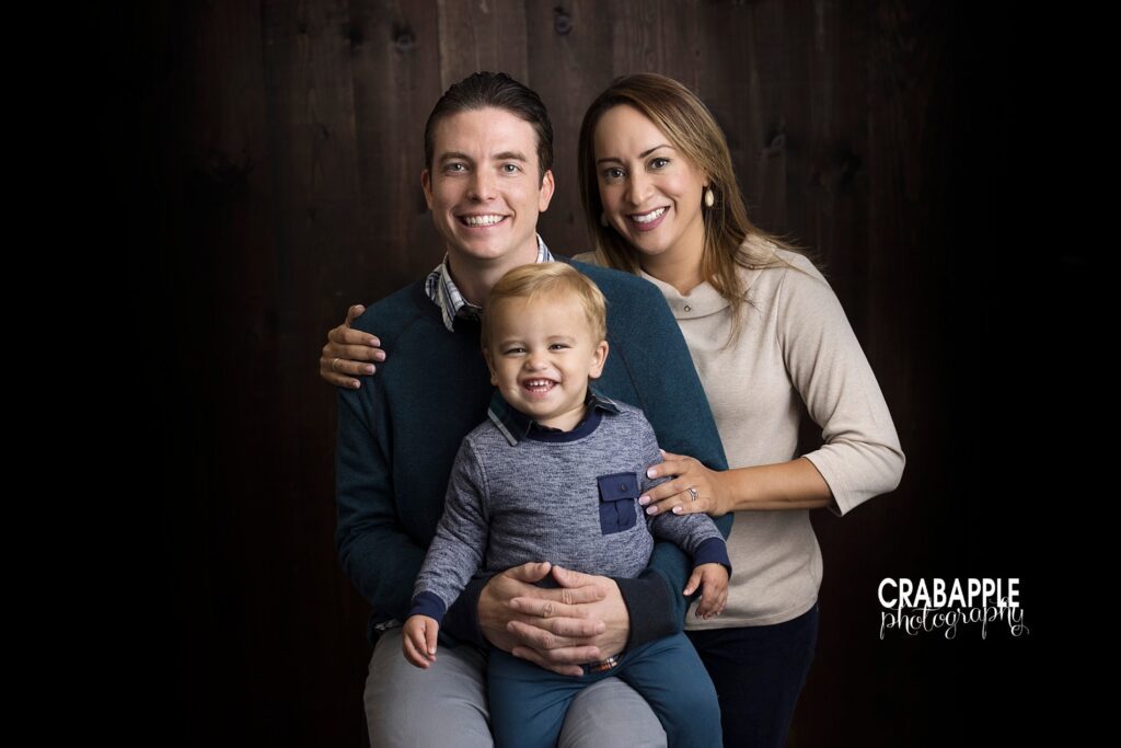 A family photo with mom, dad, and 21 month old baby son in front of a dark background. Mom is standing with her arm around dad and hand on son. Dad is sitting on a stool with his son in his lap. Everyone is smiling.