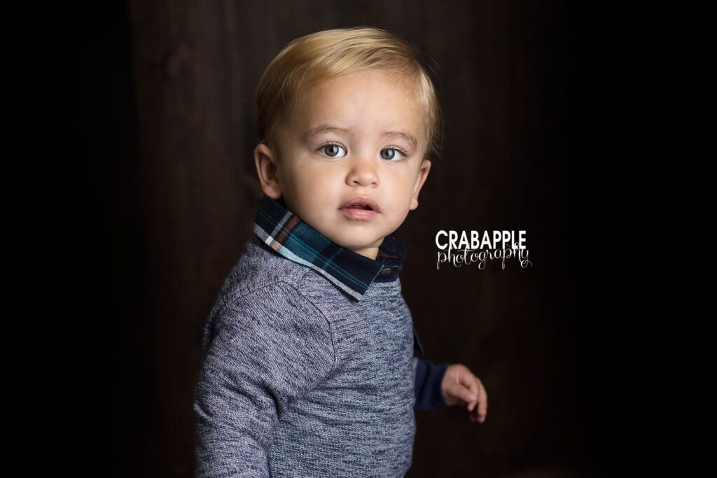 Toddler portrait of a 21 month old baby boy in front of a dark background, he is staring into the camera wearing a gray sweater over a plaid collared shirt.