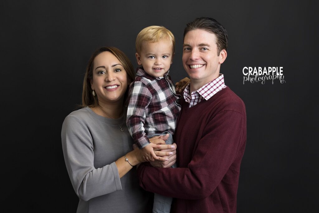 Smiling family photo of mom, dad, and son in front of a dark background. Father and son are wearing coordinating burgundy shirts and mom is in gray.