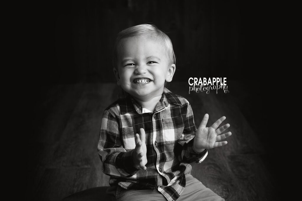 Black and white portrait of a smiling and clapping toddler boy. He is in front of a dark backdrop and wearing a plaid shirt.