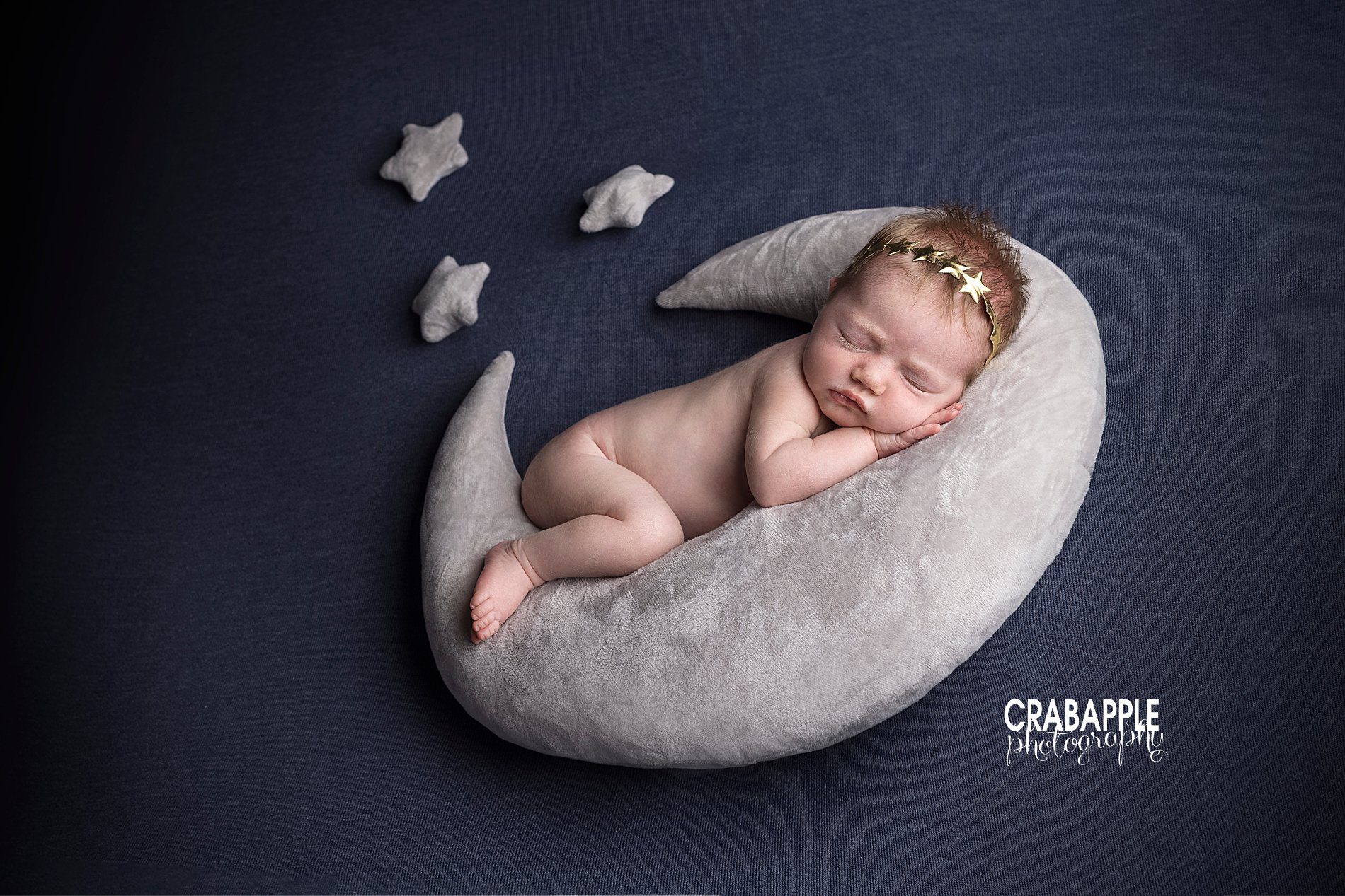 Moon themed newborn baby girl portraits using a crescent moon shaped pillow, small pillow stars, all on a navy blue blanket backdrop. Baby girl hugs the moon and wears a golden star headband.