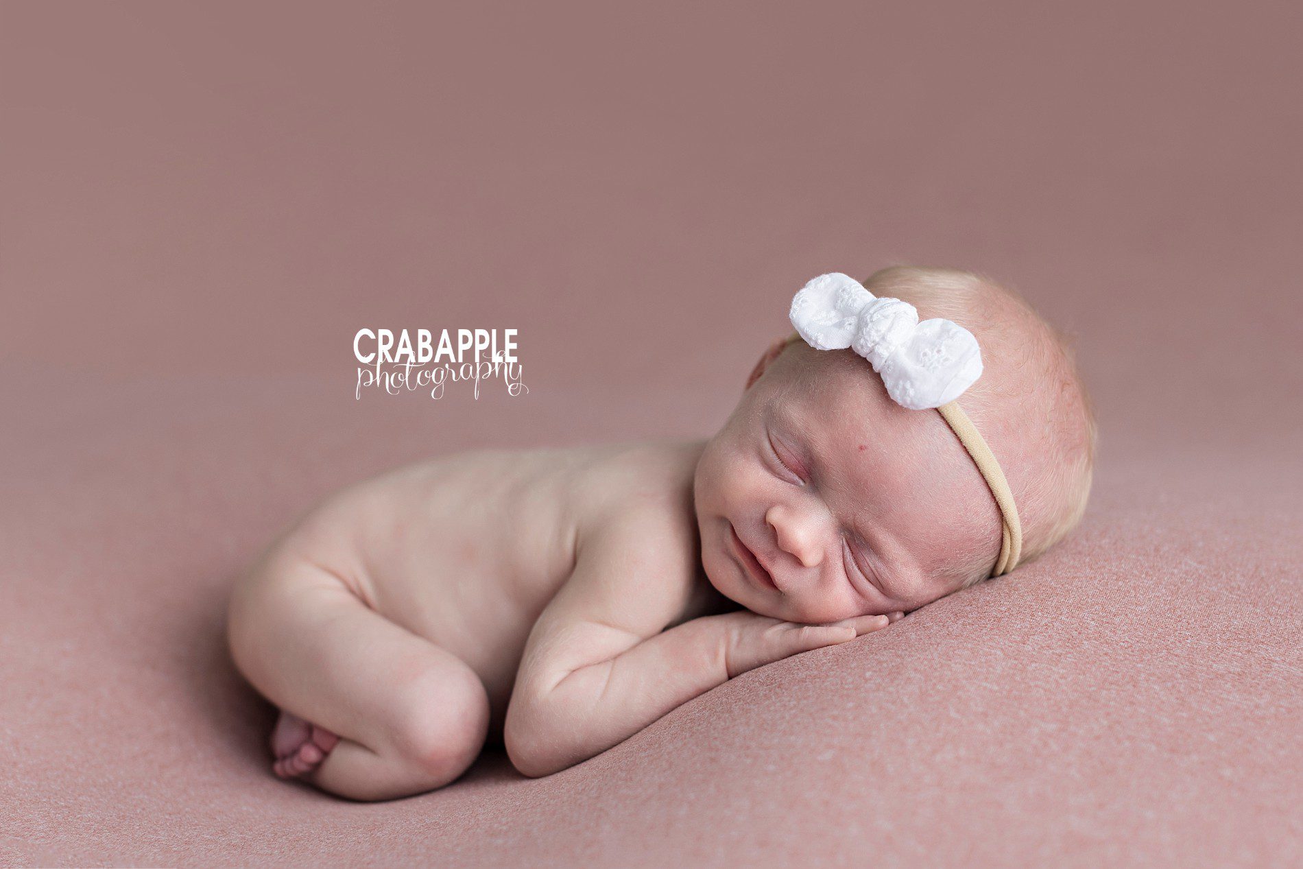 Newborn baby girl portraits with a light pink blanket as the background. Baby girl wears a white bow headband.