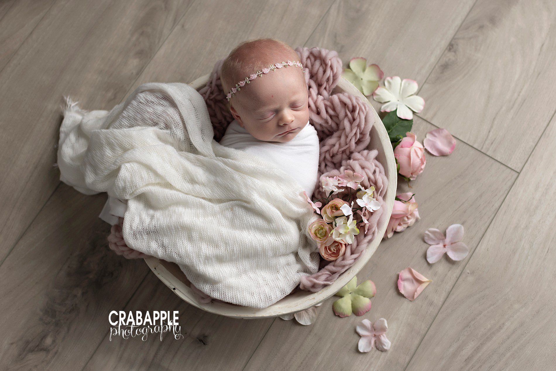 Newborn baby girl portrait of baby swaddled in white with a dusty pink knit blanket, faux flowers, and scattered faux flower petals.