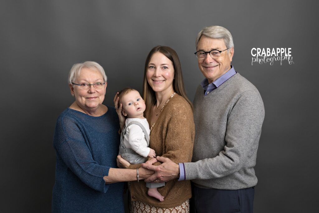 Family photo ideas with grandparents. Mom, baby, grandmother, and grandfather all stand in front of a gray backdrop and look at the camera.