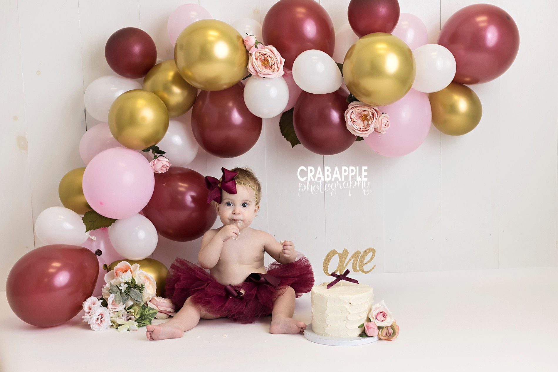 Balloon arch ideas for cake smash photos. An arch with burgundy deep red, gold, blush pink and white balloons and pink flowers.