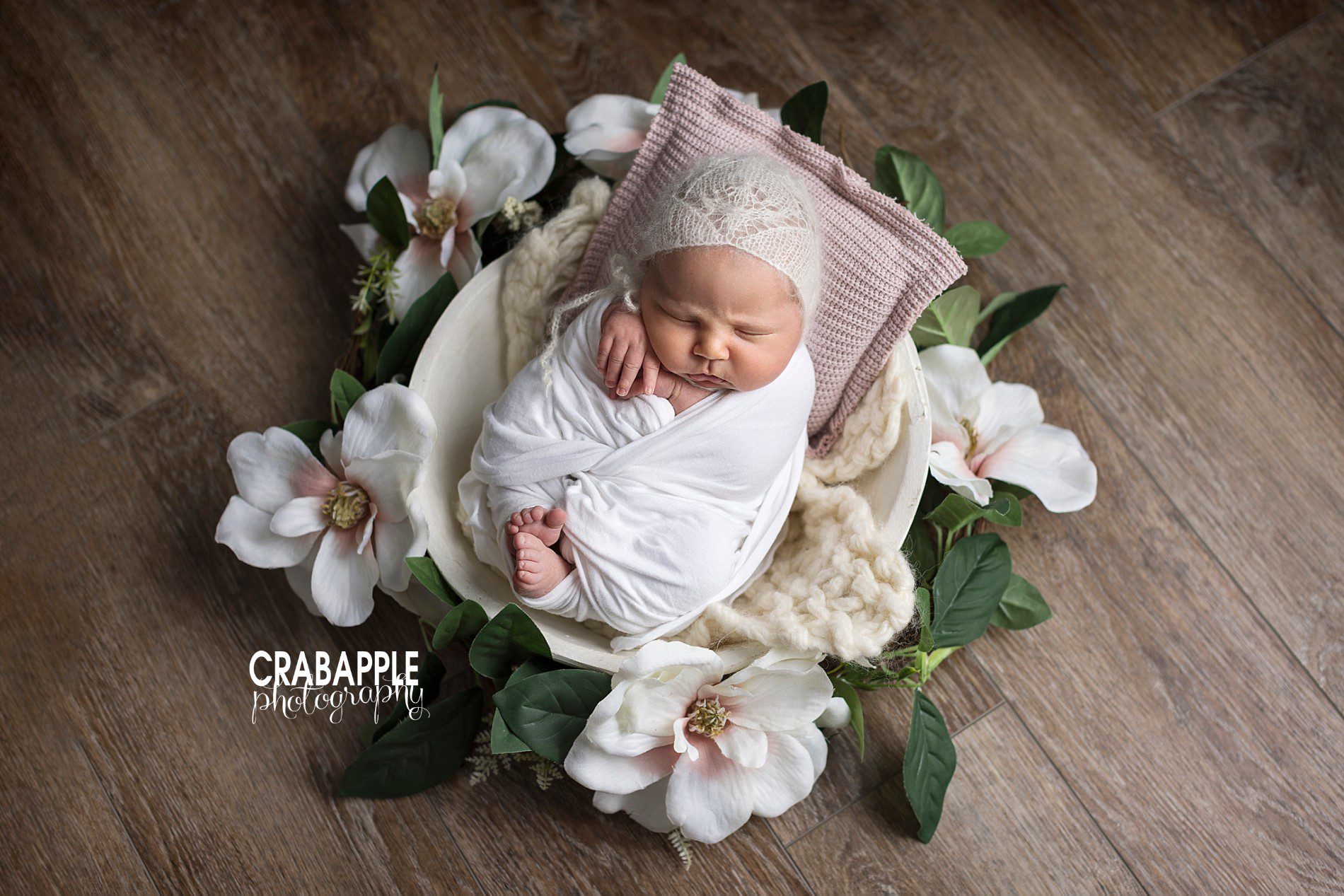 Floral newborn portraits, a baby girl swaddled in white lays in a small white bowl surrounded by faux flowers.