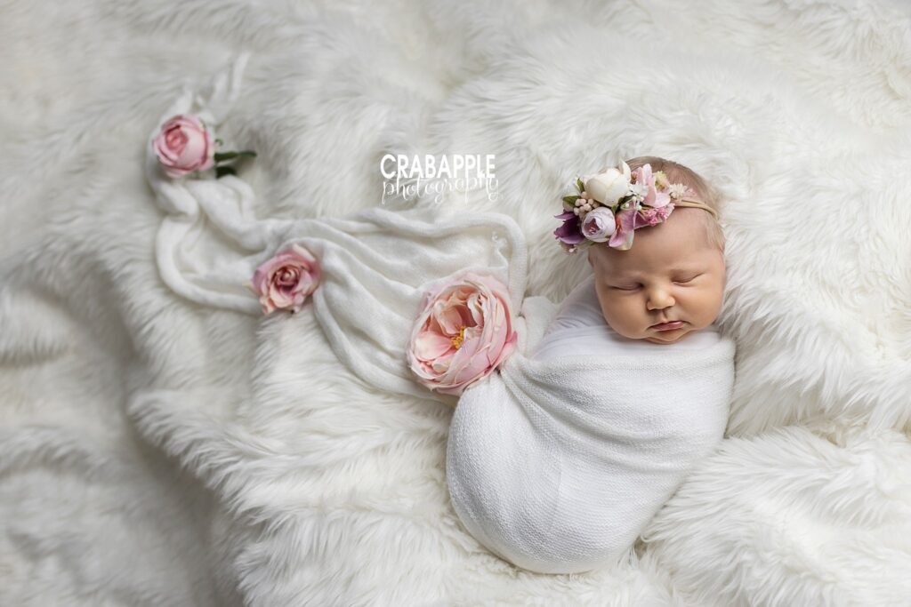 Floral newborn portraits, baby girl is swaddled in white on a white fur. Faux pink flowers scatter around her matching her floral headband.