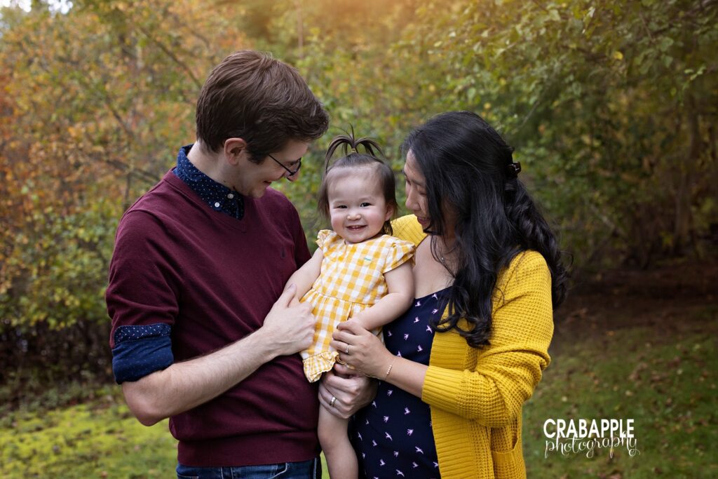 Outdoor fall family photoshoot, mom and dad hold their toddler daughter, looking at her while she looks at the camera.