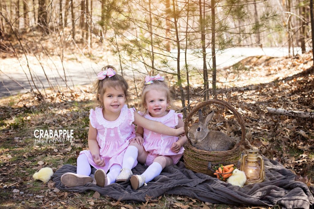 Sister photos for Easter. Two toddler girls in matching pink Easter dresses sit outside with a real bunny rabbit.