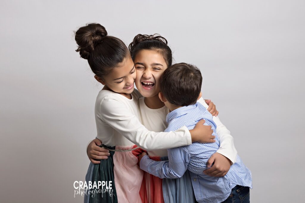 Sibling photo of three siblings ages 4-8 in a group hug in front of a light gray background.