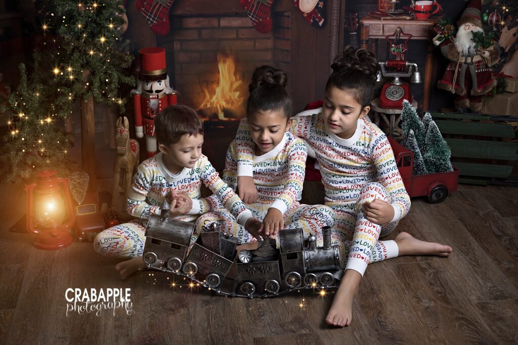 sibling portrait of three children in matching pajamas in front of a Christmas themed fireplace set playing with a vintage inspired toy train.