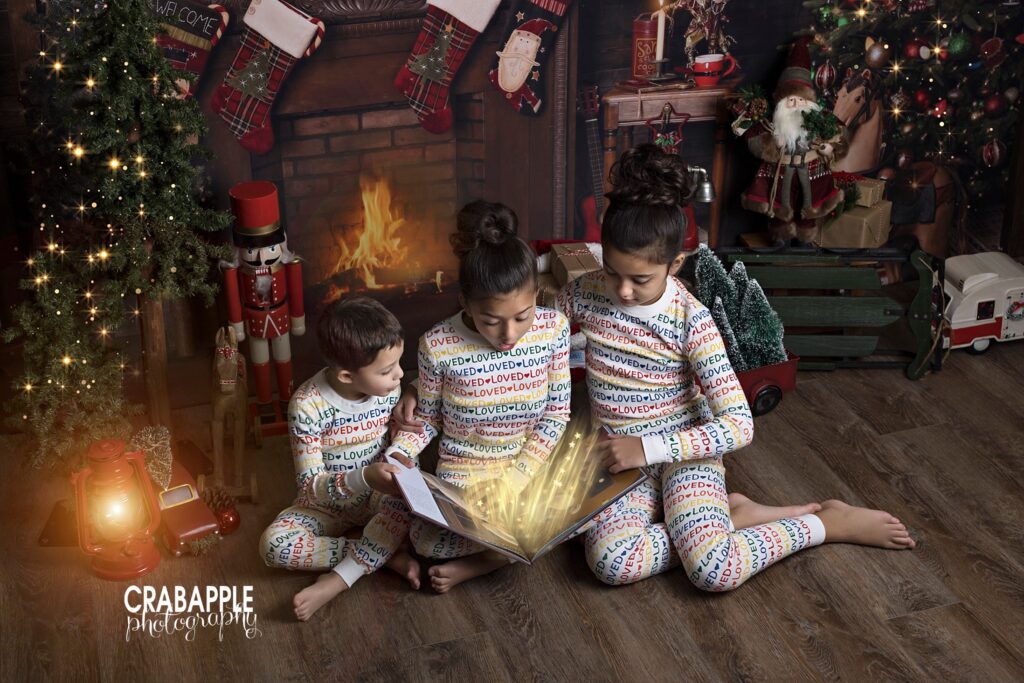 Christmas photos for siblings, three siblings in matching pajamas sit reading a Christmas book in front of a backdrop featuring a fireplace. Digital sparkles and magic are added.