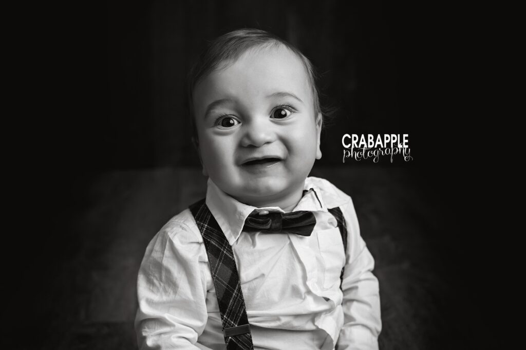 Black and white baby photo of a 1 year old boy wearing a white button down, bowtie and suspenders.