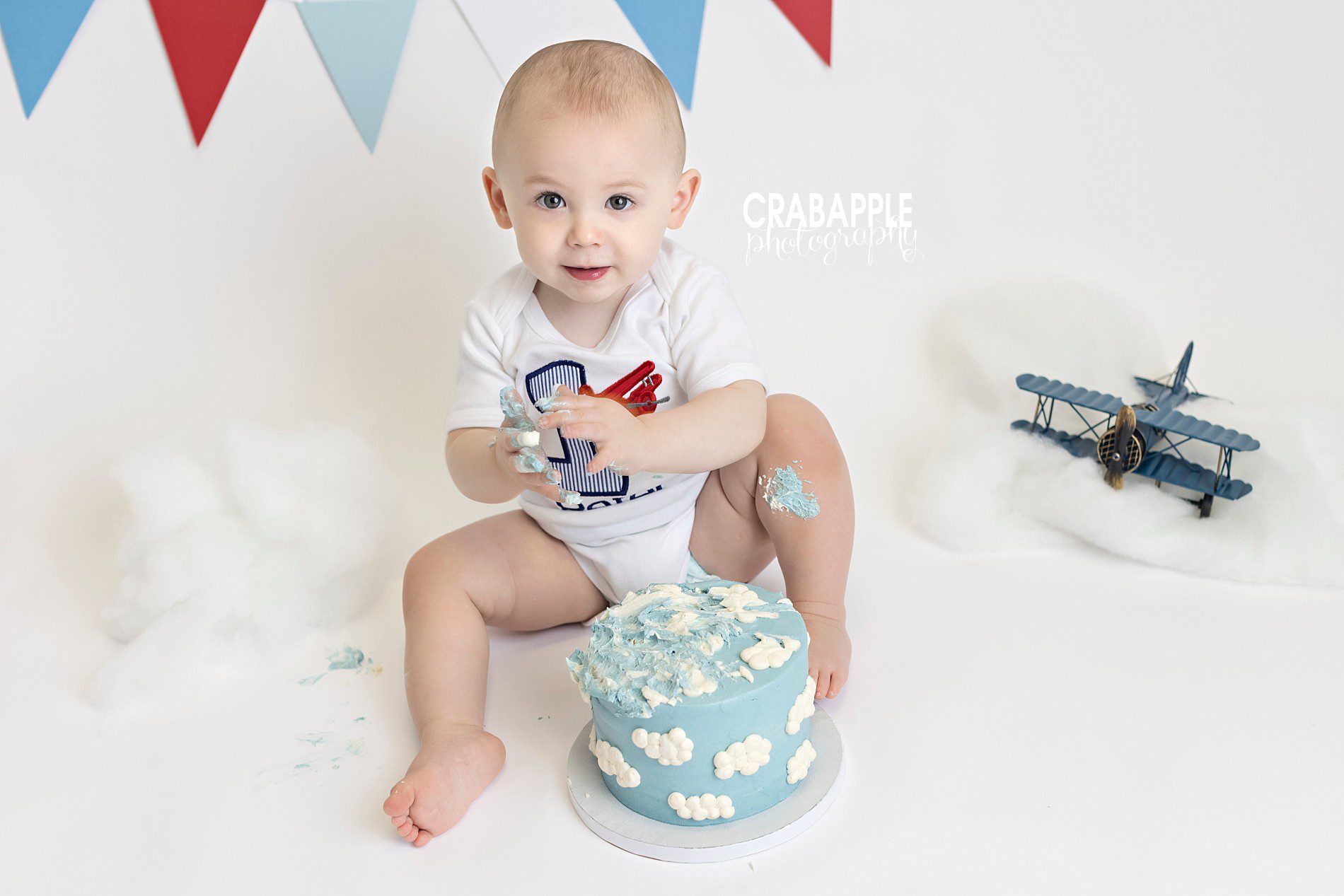 Classic plane themed cake smash with cloud cake, white background, red and blue bunting, and cotton clouds.