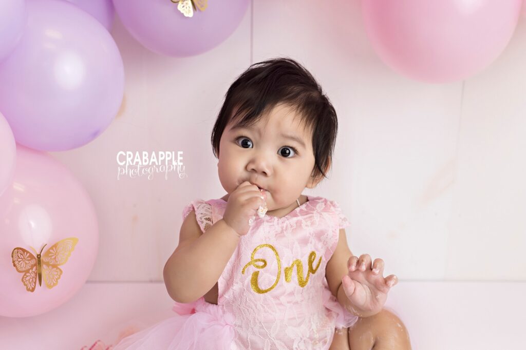 1 year old girl surrounded by light colored balloons celebrates first birthday with cake smash near Quincy MA