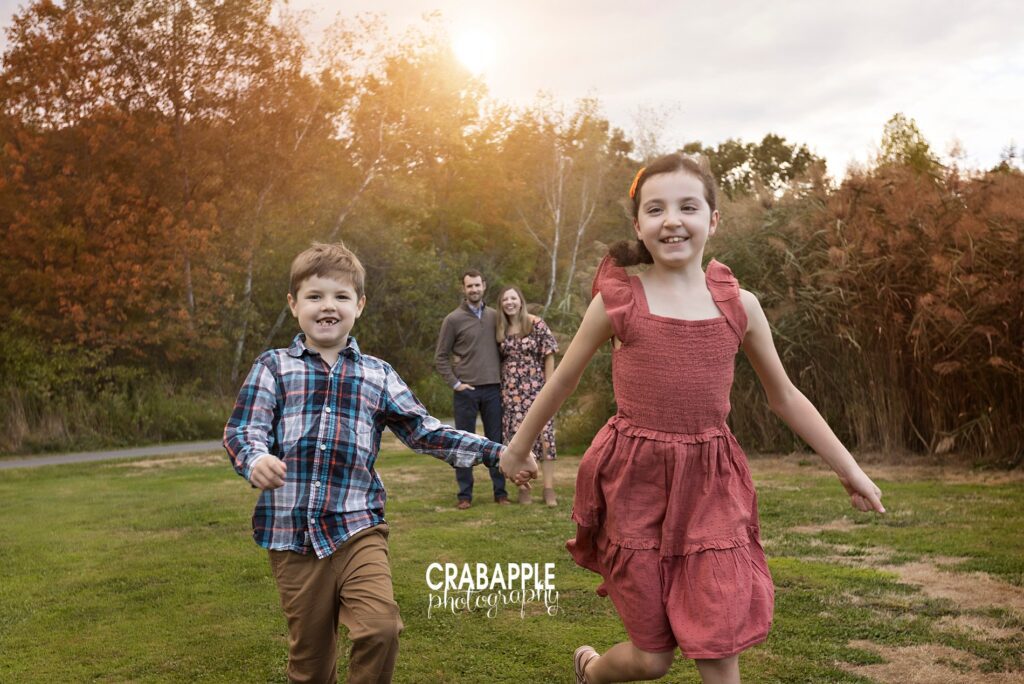 Two children hold hands and joyfully run towards the camera while the parents embrace in the background during early fall family photos
