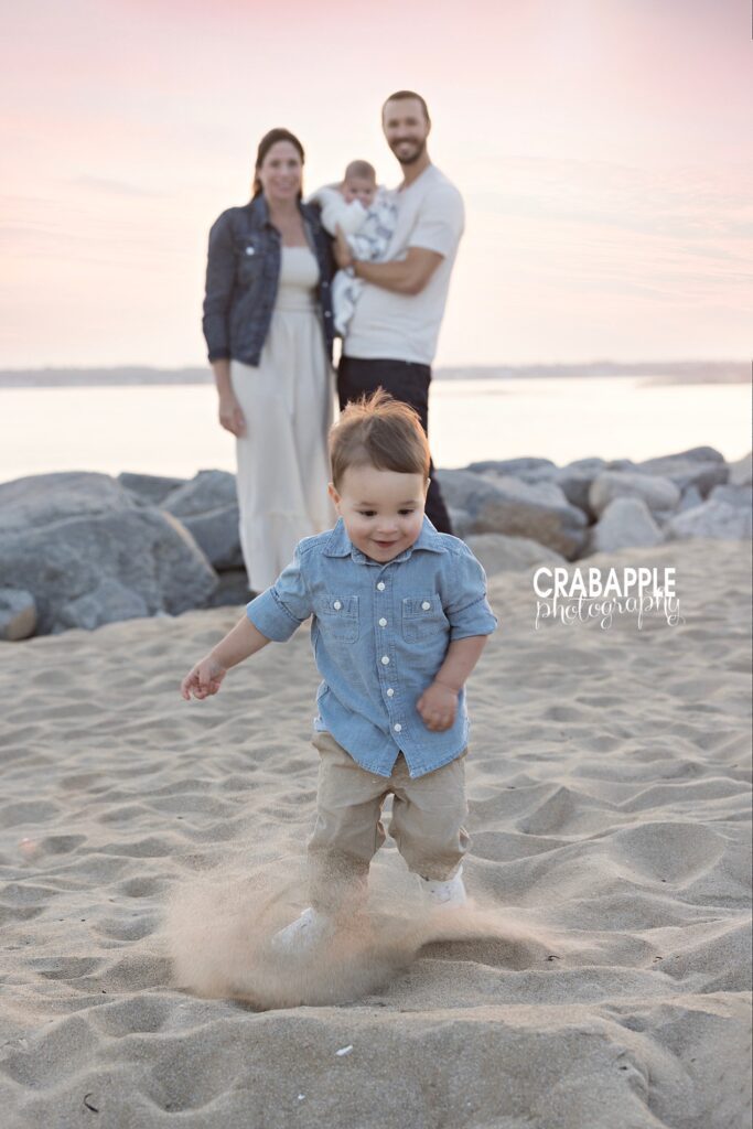 Toddler runs through the sand in the foreground while mom, dad and infant watch on in the background slightly out of focus