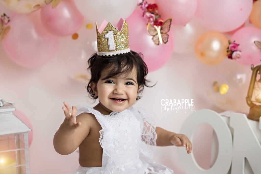 Smiling 1 year old girl in a golden  crown surrounded by balloons and butterflies during Quincy MA cake smash session