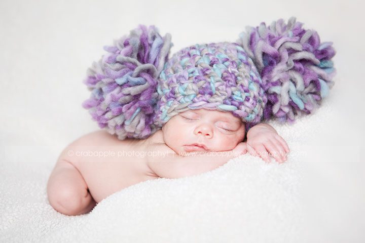 Newborn baby with oversized purple pom-pom hat sleeping on a white blanket