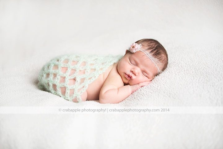 Newborn swaddled in light green loose knit wrap on white blanket with flower headband 