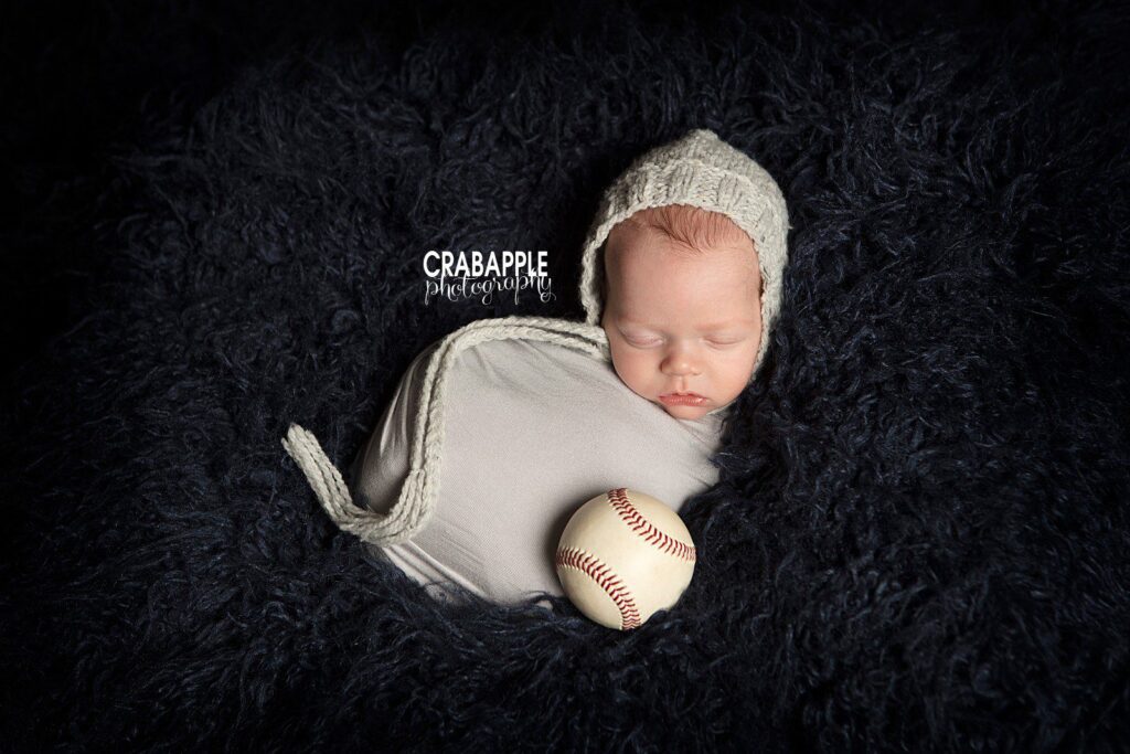 Newborn wrapped in cream blanket with baseball prop 
