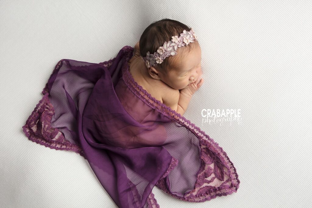 Baby girl in purple wrap with white headband on white blanket 

