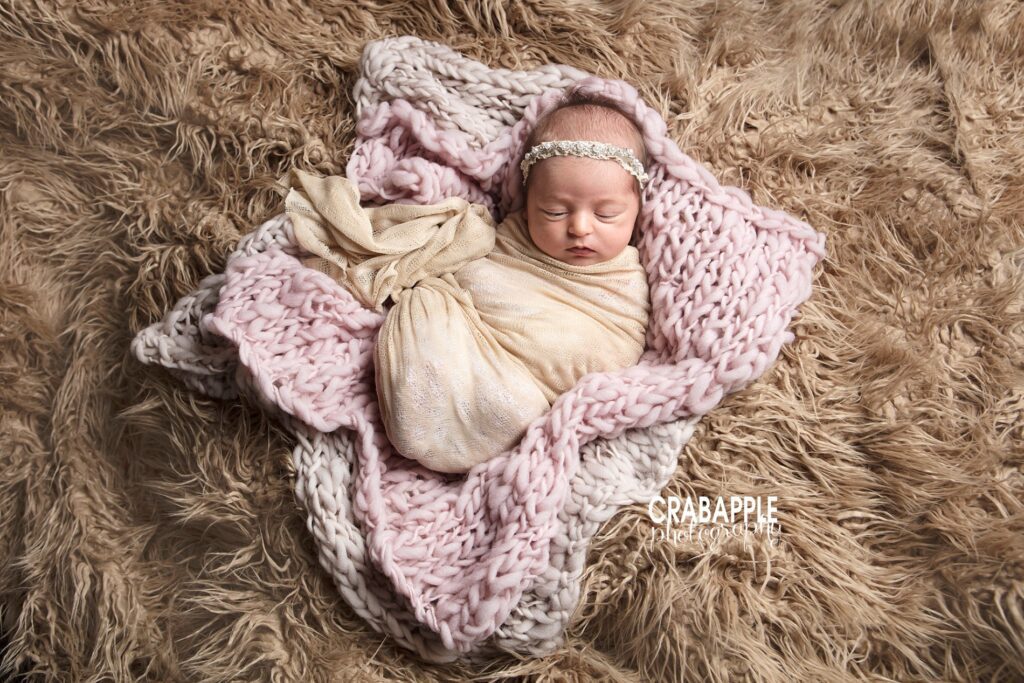 Newborn wrapped in cream and blush, lying on textured faux fur and pink knit blanket.
