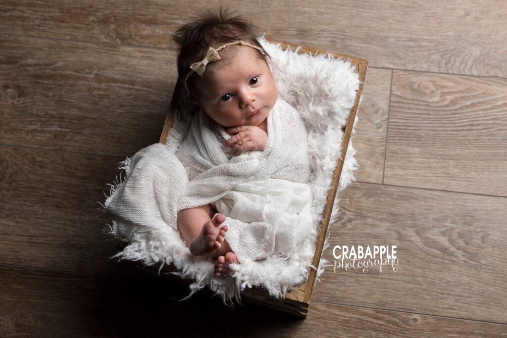 Baby girl in white wrap and headband posed in rustic wooden box on wood floor.