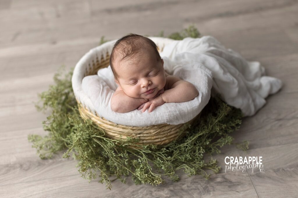 Swaddled newborn baby in moss-lined round bowl on rustic wood background.