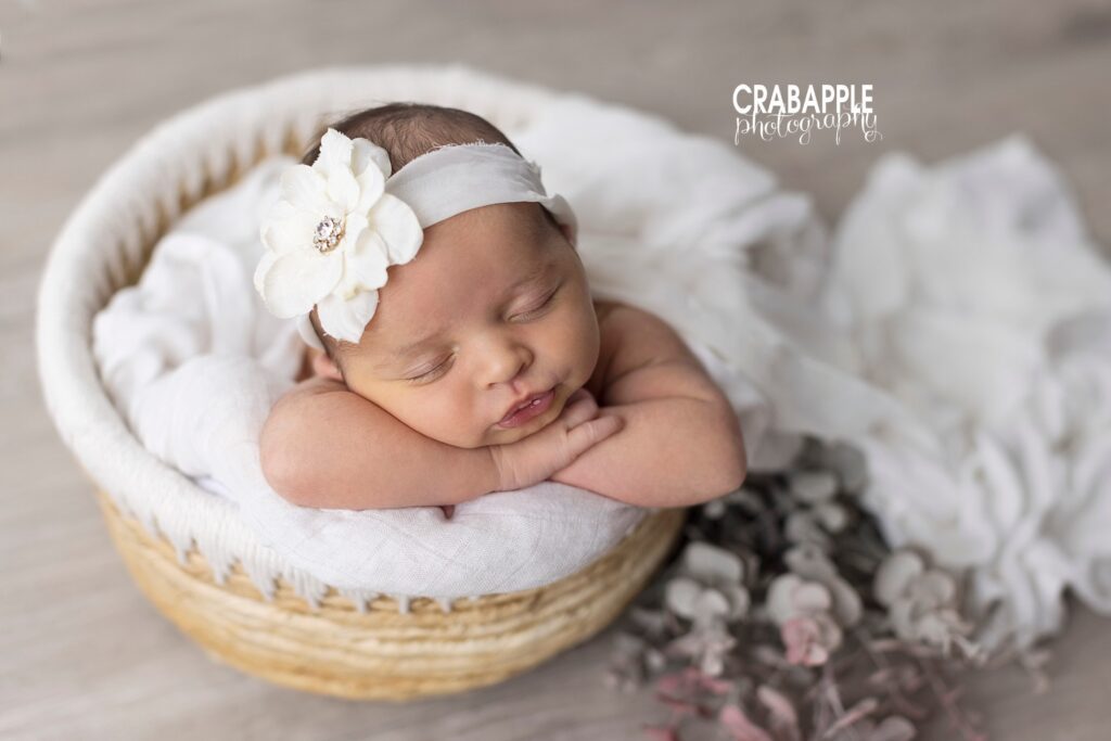 Newborn girl with floral headband posed in a basket with white blanket and soft textures.
