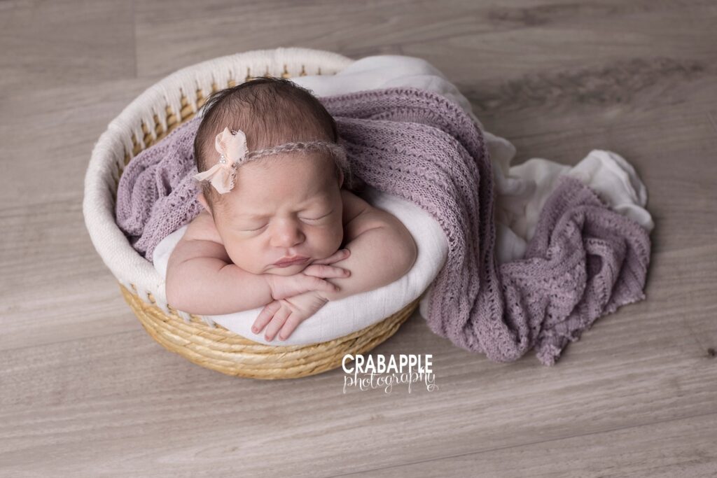 Baby girl in light purple wrap with bow headband, posed in basket with textured blankets.