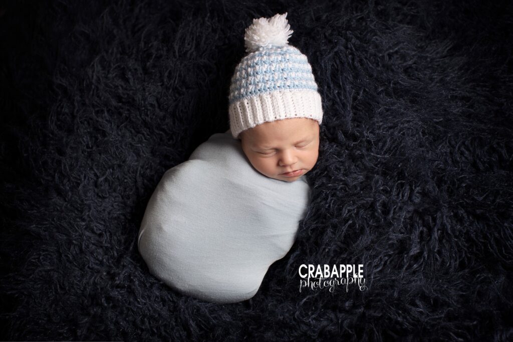 Baby boy swaddled in white with knit hat on dark blue fur 
