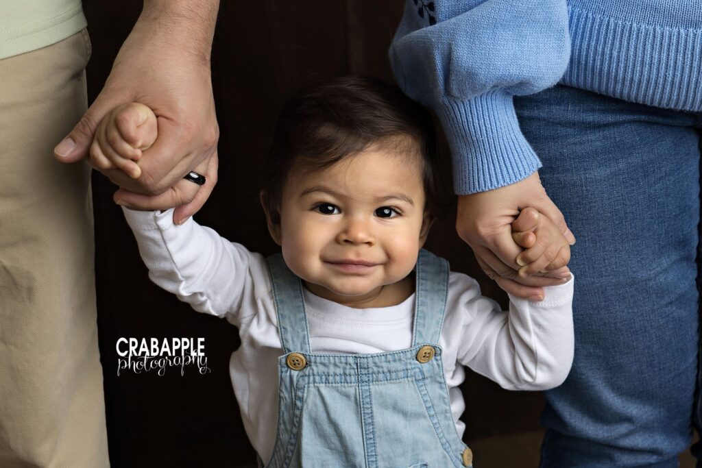 1 Year old boy smiles for camera while holding hands with mom and dad
