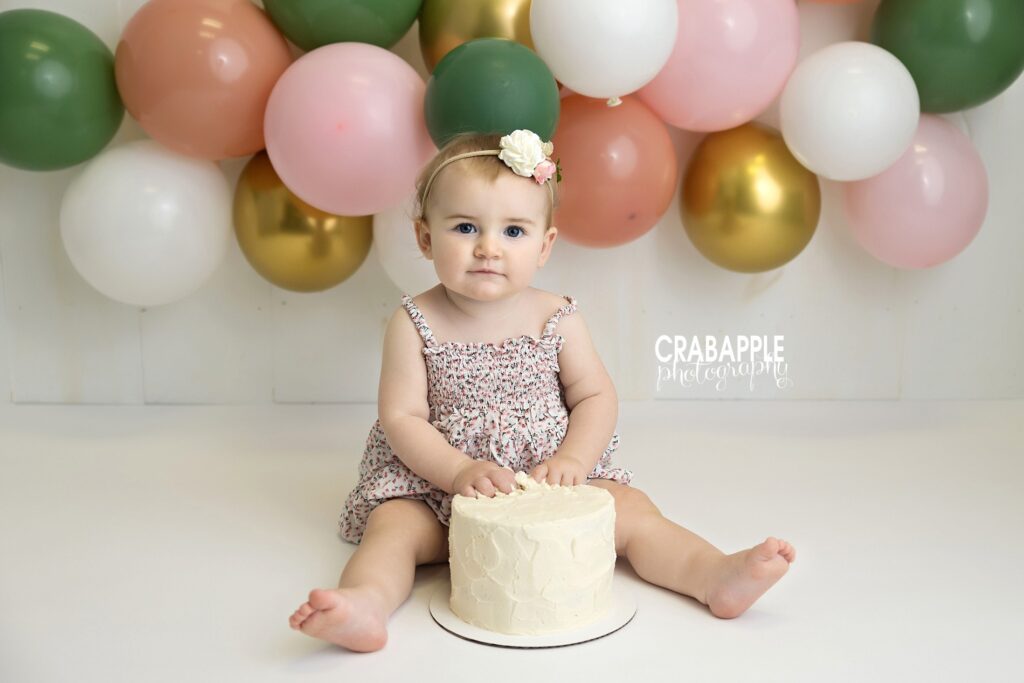 One-year-old baby smiling gently while surrounded by bright, colorful balloons, white cake, with a clean face.