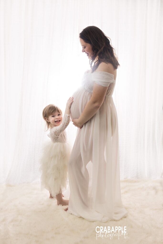 Ethereal portrait of pregnant mother in flowy white gown and toddler daughter in matching tutu, with daughter embracing the baby bump