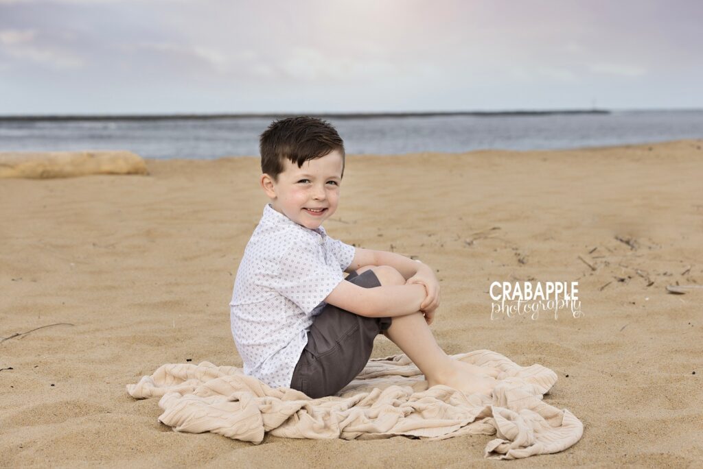Young boy sitting on a blanket in the sand during beach family portraits a perfect example of childhood at the beach by a family photographer near Plum Island