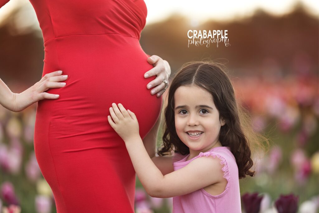Close up photo of expectant mother in a form fitting red gown with toddler older sister embracing the belly and smiling at the camera