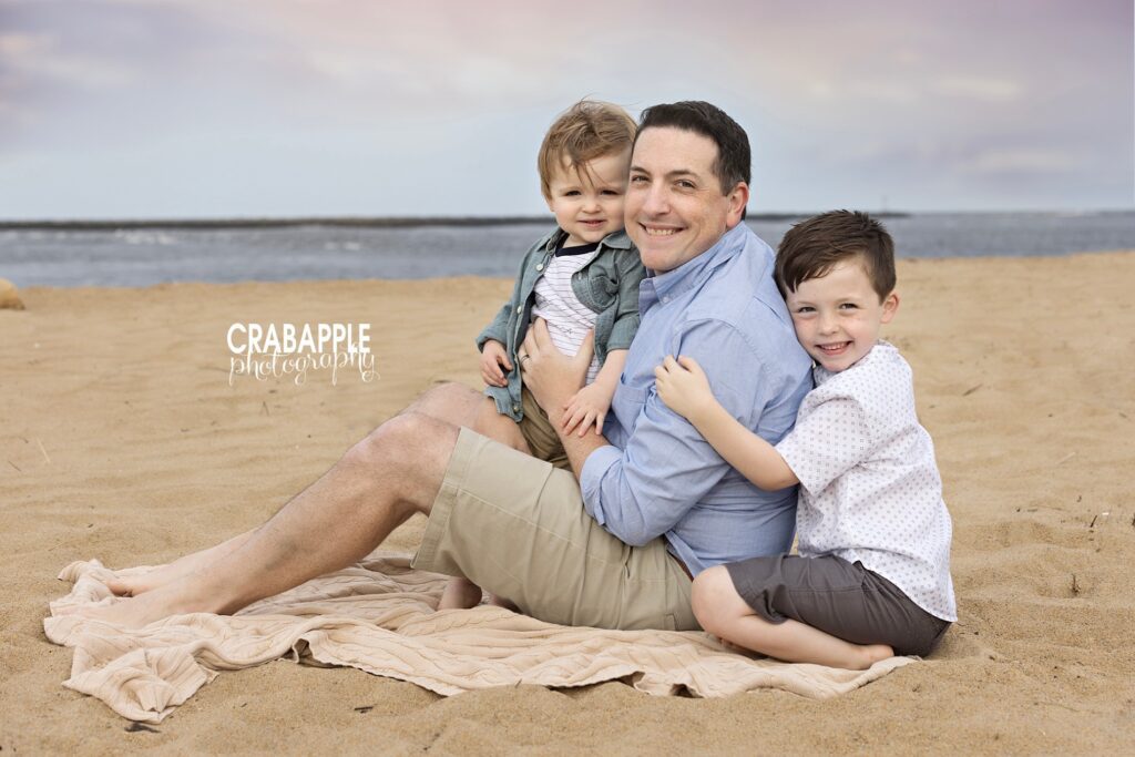 Father sitting in the sand with two sons during beach family session near Boston