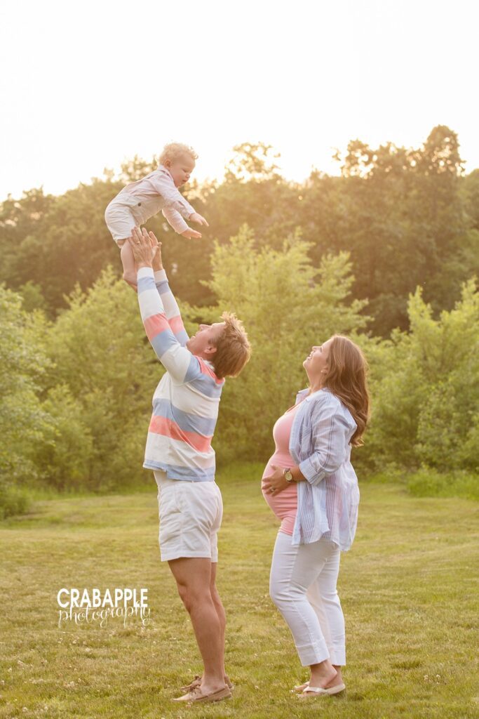 A dad playfully tosses older son up in the air while mom watches and smiles during an outdoor Southern New Hampshire maternity photo session