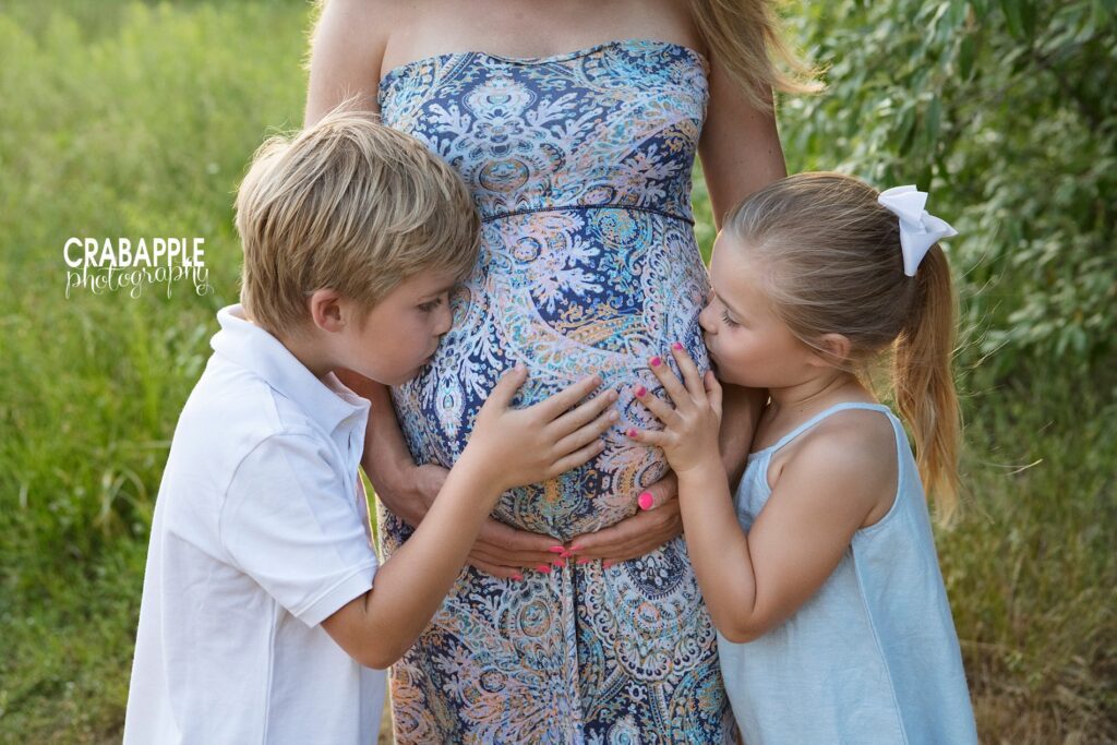 Young brother and sister hugging mom’s baby bump outdoors