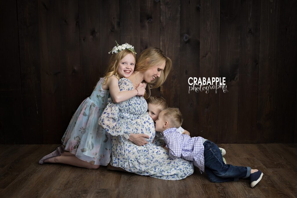 Siblings sitting together hugging mom and kissing belly during studio maternity photo